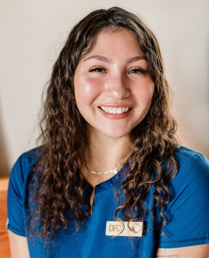 Chiropractic assistant smiling in blue uniform with "DPC" logo, representing the caring team at Desert Pine Chiropractic in Bellingham.