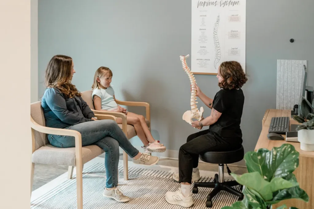 Woman explaining spinal health to a child and her mother during a chiropractic consultation, with a spinal model and educational poster in a modern clinic setting.