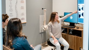 Chiropractor explaining spinal health to patients in consultation room with anatomical spine model and x-ray images on screen.