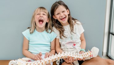 Two smiling children holding a model spine, representing pediatric chiropractic care and the importance of spinal health at Desert Pine Chiropractic in Bellingham, WA.